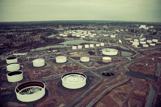 A complex network of pipelines and storage tanks at a large oil refinery, with a faint haze over the industrial landscape, suggesting high operational activity but also potential environmental impact.