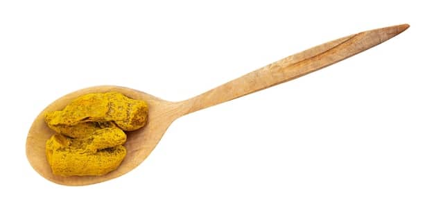 A wooden spoon scooping a spoonful of vibrant yellow turmeric powder from a small bowl, highlighting its rich color and fine texture. The bowl sits on a rustic wooden table, with whole turmeric roots and other spices scattered around, emphasizing the spice's natural and potent anti-inflammatory properties.
